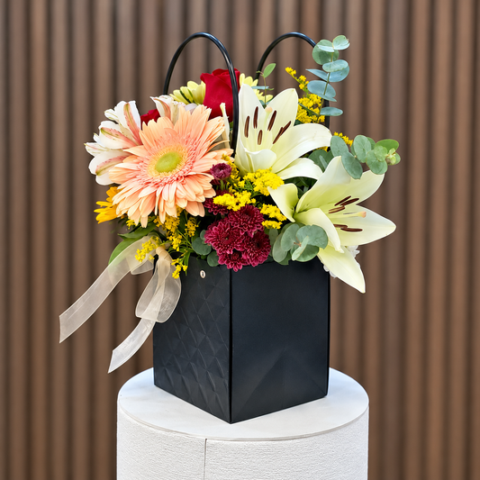 Arreglo en bolso negro con lirios blancos, gerbera durazno y lazo beige, vista lateral. Velve Floristería, Paseo de las Flores, Heredia.