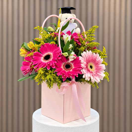 Cartera rosada con gerberas fucsias y rosadas, astromelias, solidago y osito de graduación, vista diagonal. Velve Floristería, Paseo de las Flores, Heredia.