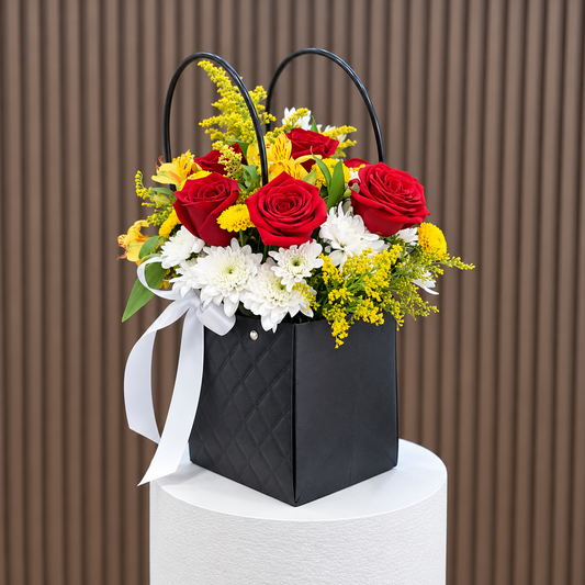 Vista diagonal de canasta negra con rosas rojas, pomas blancas, astromelias amarillas, solidago y lazo blanco sobre pedestal blanco. Velve Floristería, Paseo de las Flores, Heredia.