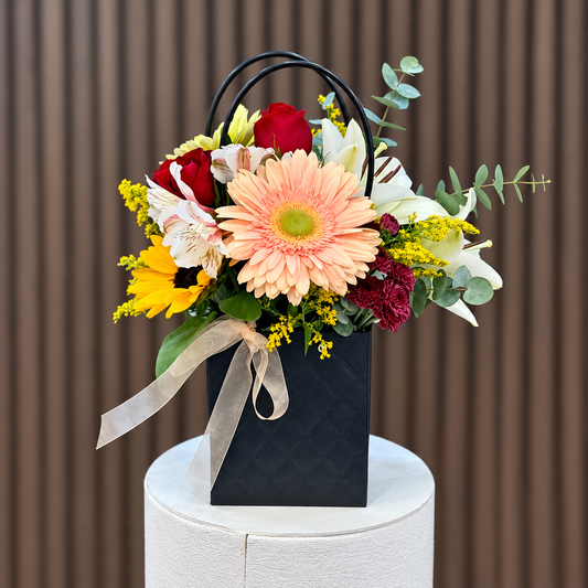 Arreglo en bolso negro con gerbera durazno, rosas rojas, lirios blancos y eucalipto, vista frontal. Velve Floristería, Paseo de las Flores, Heredia.