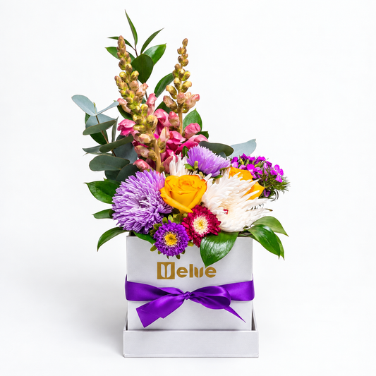 Arreglo de rosas amarillas, crisantemos blancos y lilas, flores rosadas y eucalipto en caja blanca con lazo morado y logo dorado de Velve, visto de lado. Velve Floristería, Paseo de las Flores, Heredia.