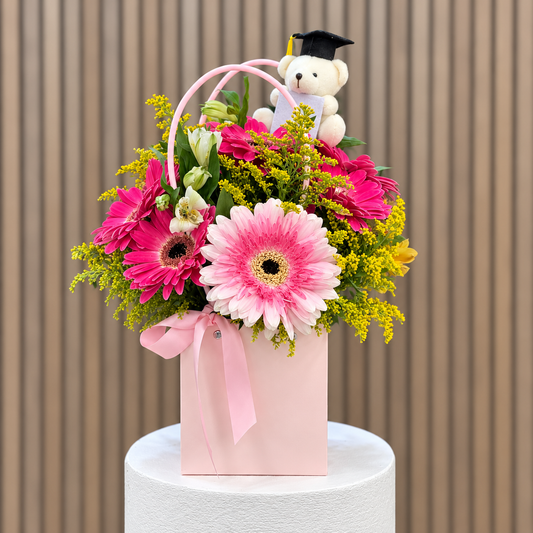 Cartera rosada con gerberas, astromelias, solidago y osito de graduación, vista frontal. Velve Floristería, Paseo de las Flores, Heredia.