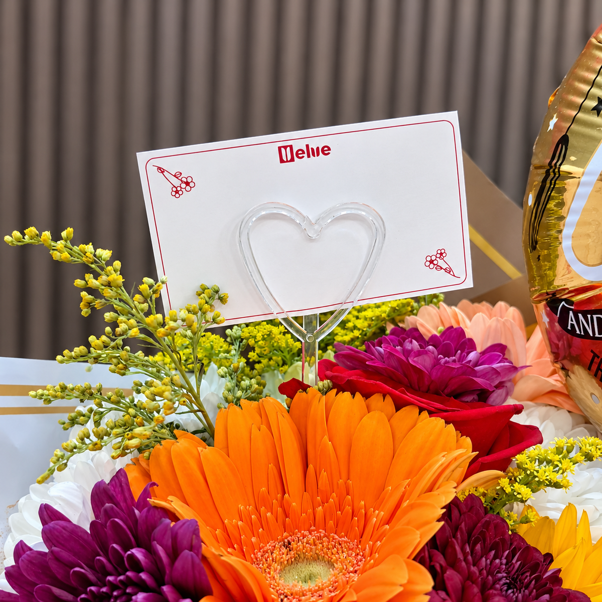 Detalle de tarjeta para dedicatoria con logo de Velve y pincho transparente de corazón entre gerbera naranja, crisantemos morados y globo dorado. Velve Floristería, Paseo de las Flores, Heredia.