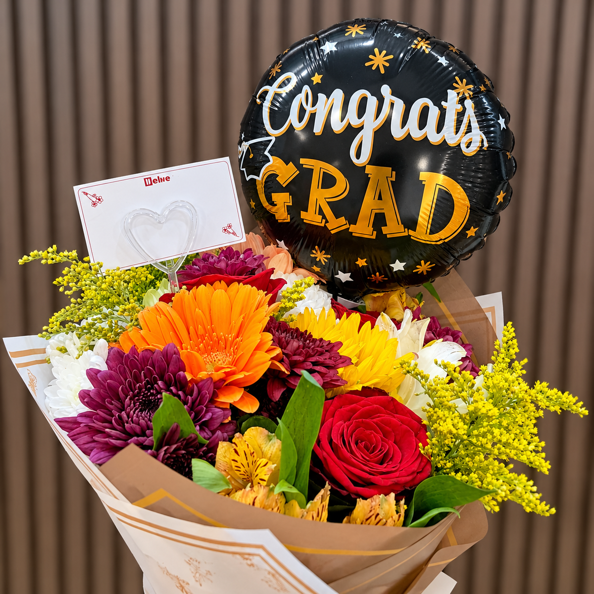 Ramo mixto de graduación con gerbera naranja, rosas rojas, girasol, crisantemos y solidago, acompañado de globo dorado «Congrats Grad» y tarjeta para dedicatoria. Velve Floristería, Paseo de las Flores, Heredia.