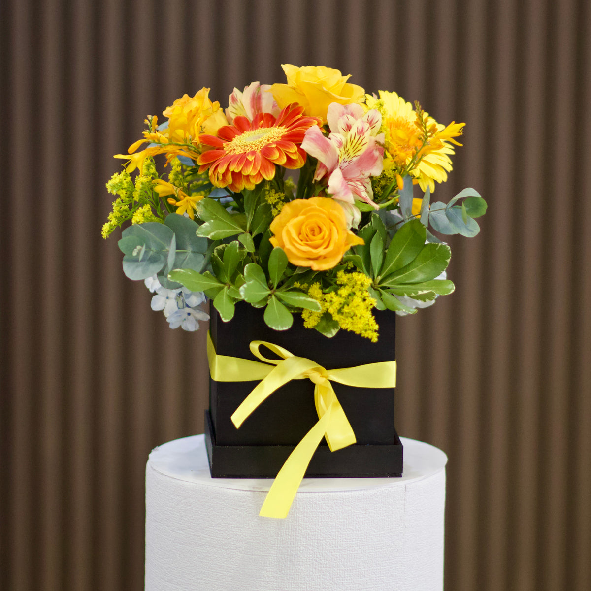 Vista frontal de arreglo en caja de madera negra con rosas amarillas, gerberas amarilla y anaranjada, astromelias rosadas, follaje verde y lazo amarillo. Velve Floristería, Paseo de las Flores, Heredia