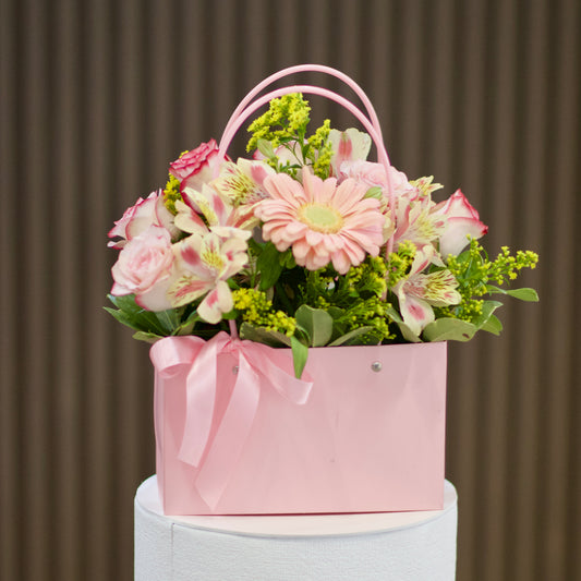 Vista frontal de cartera rosada con gerbera y rosas en tonos rosados, astromelias, solidago amarillo y follaje verde, decorada con lazo rosa. Velve Floristería, Paseo de las Flores, Heredia