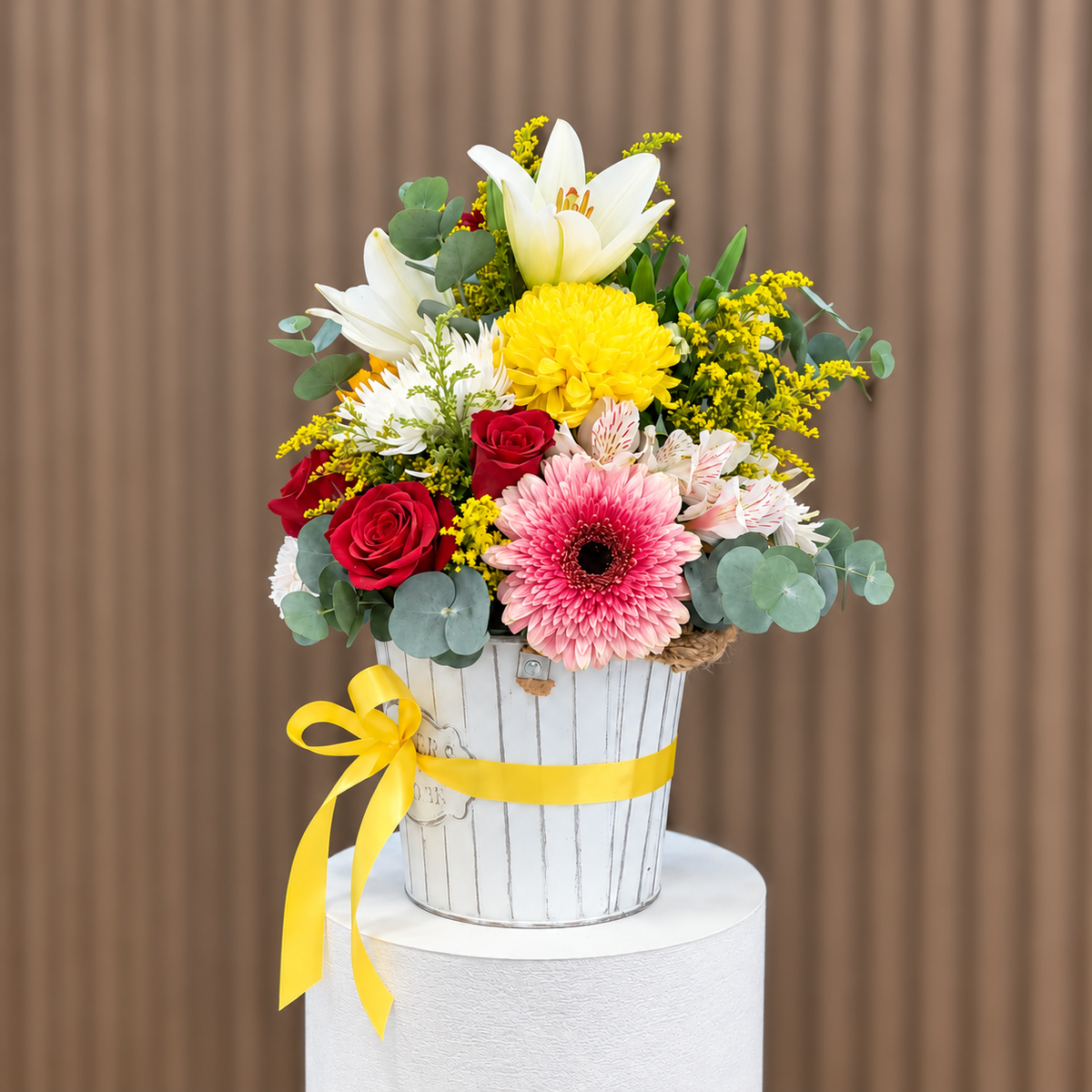 Vista diagonal de arreglo con rosas rojas, gerbera rosada, lirios blancos, crisantemo amarillo y astromelias en base blanca con lazo amarillo. Velve Floristería, Paseo de las Flores, Heredia.