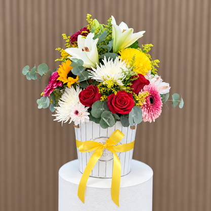 Vista frontal de arreglo con rosas rojas, lirios blancos, gerberas rosadas, girasol y eucalipto en base blanca con lazo amarillo. Velve Floristería, Paseo de las Flores, Heredia.