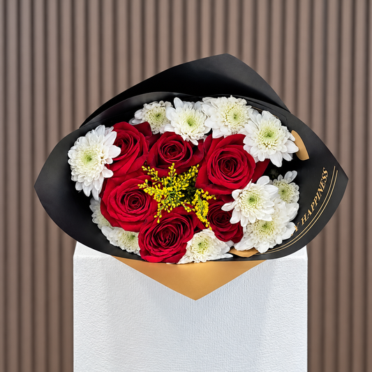 Vista frontal de ramo de seis rosas rojas con pomas blancas y solidago en envoltura negra y dorada sobre pedestal blanco. Velve Floristería, Paseo de las Flores, Heredia.