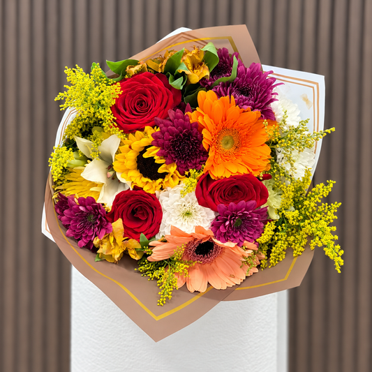 Vista frontal de ramo mixto con rosas rojas, girasol, gerberas naranja y durazno, lirio blanco y crisantemos en envoltura blanca y café con bordes dorados. Velve Floristería, Paseo de las Flores, Heredia.