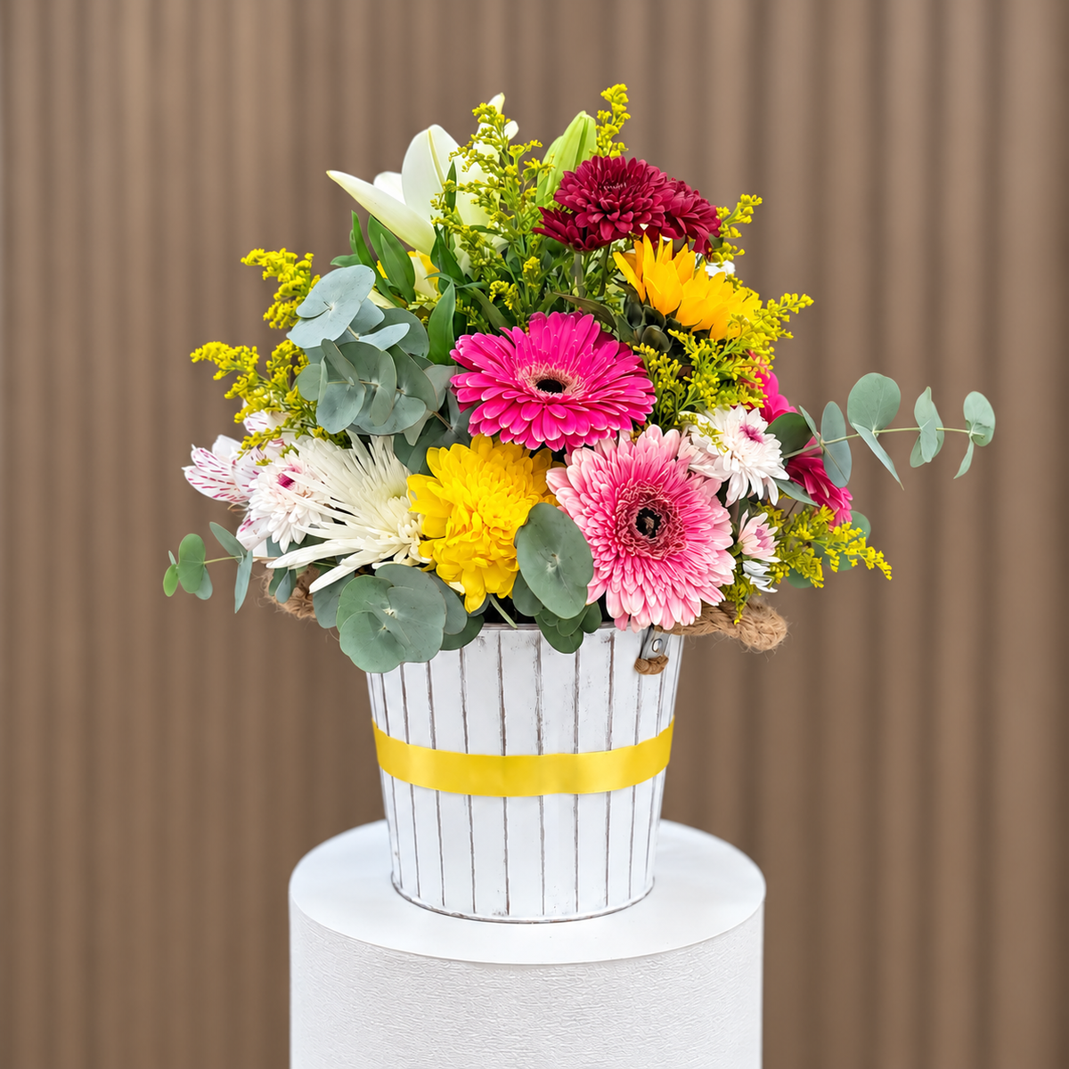 Vista posterior de arreglo con gerberas rosadas, girasol, lirio blanco, crisantemos y eucalipto en base blanca con cinta amarilla. Velve Floristería, Paseo de las Flores, Heredia.