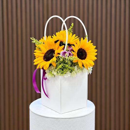 Arreglo de girasoles y flores moradas en bolso blanco, vista diagonal. Velve Floristería, Paseo de las Flores, Heredia.