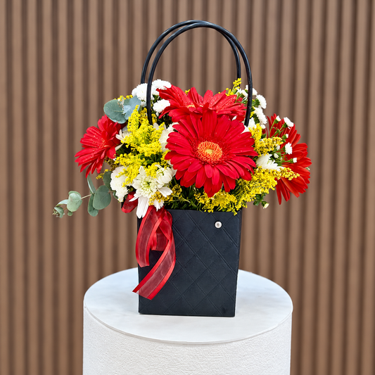 Cartera negra con gerberas rojas, flores blancas, eucalipto y lazo rojo, vista frontal. Velve Floristería, Paseo de las Flores, Heredia.