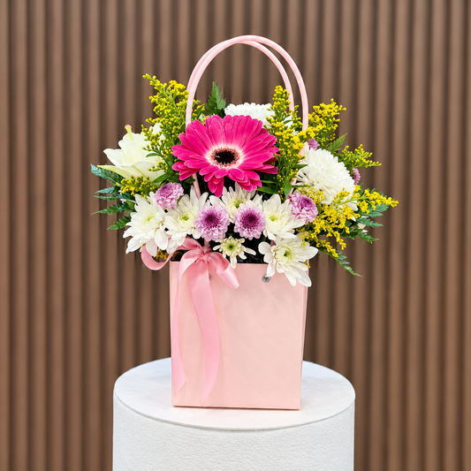 Cartera rosada con gerbera fucsia, lirio blanco, crisantemos blancos y violetas y lazo rosado, vista frontal. Velve Floristería, Paseo de las Flores, Heredia.