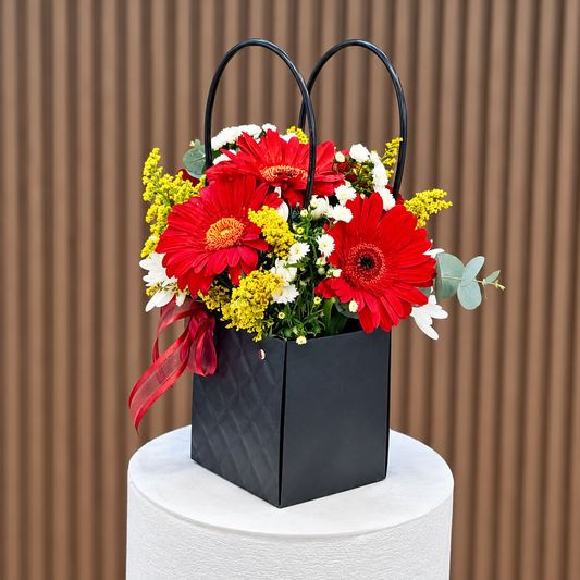 Vista lateral de cartera negra con gerberas rojas, flores blancas, eucalipto y lazo rojo. Velve Floristería, Paseo de las Flores, Heredia.