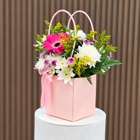 Vista lateral de cartera rosada con gerbera fucsia, crisantemos blancos y violetas y follaje. Velve Floristería, Paseo de las Flores, Heredia.
