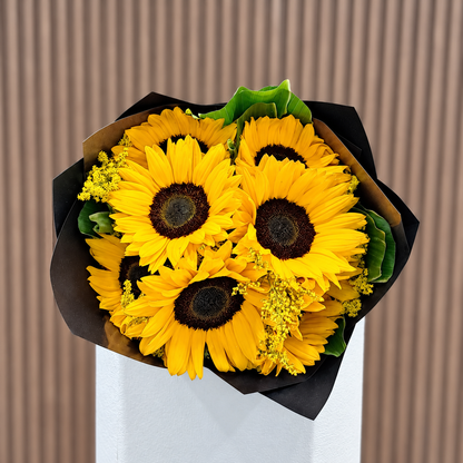Vista superior de ramo de girasoles con solidago y hojas verdes en envoltura negra y café. Velve Floristería, Paseo de las Flores, Heredia.
